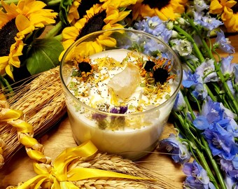 May include: A clear glass candle filled with white wax, adorned with dried flowers, a crystal, and seeds. The candle is surrounded by sunflowers, blue delphiniums, and wheat stalks, creating a natural, floral arrangement. The candle is on a wooden surface.