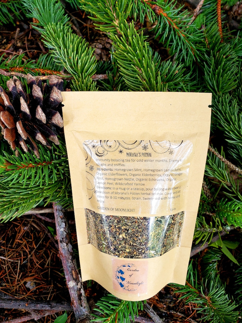 May include: A brown paper bag with a white label that says "Morana's Potion" and "Garden of Moonlight". The label also lists the ingredients for an immunity boosting tea, including mint, lemon balm, elderflowers, elderberries, ginger root, nettle, echinacea, lemon peel, and yarrow.