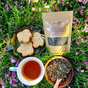 May include: A still life featuring a cup of tea, leaf-shaped cookies, and a bag of herbal tea on a bed of grass and pink flower petals. The tea is in a white cup with a floral design. The bag reads "Lada's Blessing".