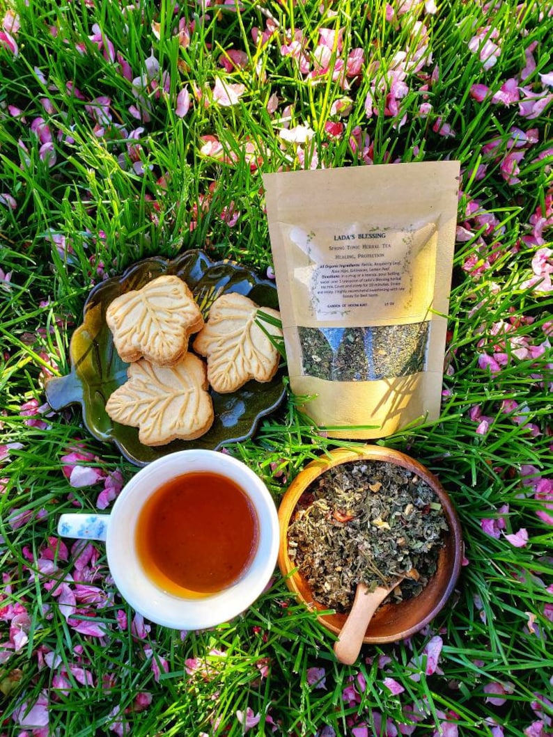 May include: A still life featuring a cup of tea, leaf-shaped cookies, and a bag of herbal tea on a bed of grass and pink flower petals. The tea is in a white cup with a floral design. The bag reads "Lada's Blessing".