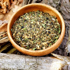 May include: A wooden bowl filled with a blend of dried herbs and flowers, including green tea, dried apple pieces, and blueberries. The bowl is sitting on a piece of weathered wood.