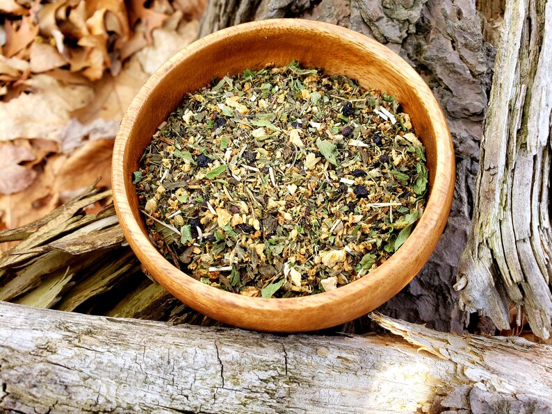 May include: A wooden bowl filled with a blend of dried herbs and flowers, including green tea, dried apple pieces, and blueberries. The bowl is sitting on a piece of weathered wood.