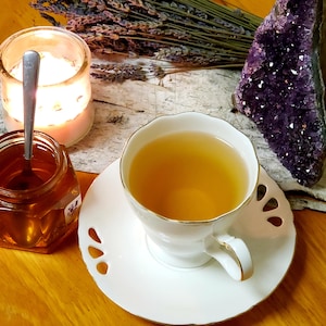 May include: A white teacup and saucer with a golden rim, filled with light yellow tea. The teacup is on a wooden surface with a purple amethyst crystal, a bunch of lavender, and a glass candle holder.