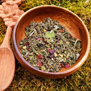 May include: A wooden bowl filled with a mix of dried herbs and berries. The bowl sits on a bed of green moss, with a wooden spoon and carved floral design visible. The herbs and berries are a mix of green, red, and brown.