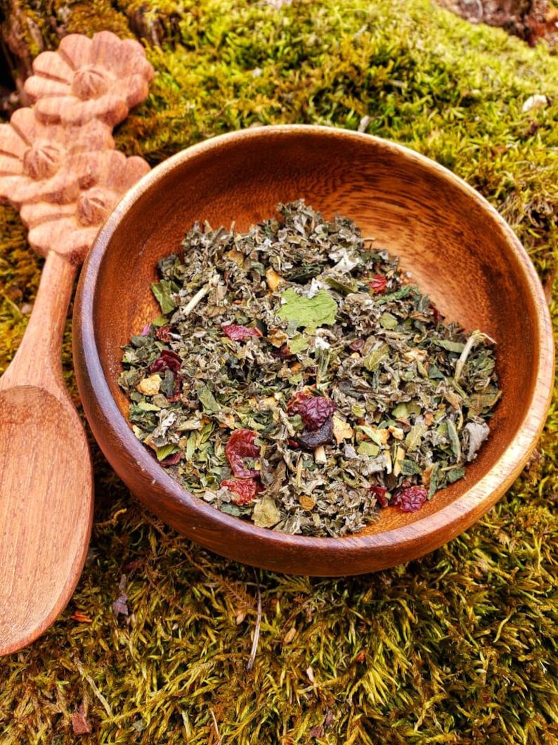 May include: A wooden bowl filled with a mix of dried herbs and berries. The bowl sits on a bed of green moss, with a wooden spoon and carved floral design visible. The herbs and berries are a mix of green, red, and brown.