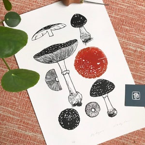 May include: A black and white mushroom print with a red mushroom cap. The print includes various mushroom illustrations and the text "Fly Agaric". A wood carving tool is placed above the print.