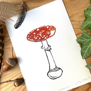 May include: A red and white mushroom with white spots on the cap and a black stem. The mushroom is drawn in a simple, stylized way. The image is signed "Amanda Rosema" and numbered "016".