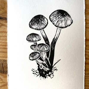 May include: A black and white linocut print of mushrooms. The print features several mushrooms with detailed caps and stems, clustered together. The artwork is on textured, off-white paper. The artist's signature and the words "Honey Fungus" are visible.