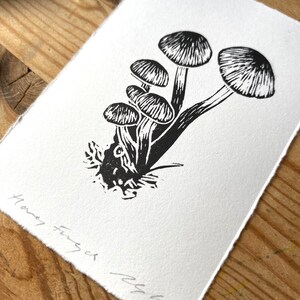 May include: A black and white linocut print of honey fungus mushrooms. The print features six mushrooms with detailed caps and stems, set against a white background. The words "Honey Fungus" and an artist's signature are visible in the lower left corner.