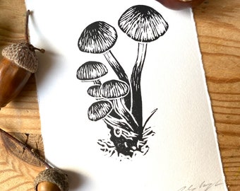 May include: Black and white linocut print of mushrooms on textured white paper. The print features several mushrooms with detailed caps and stems. The artwork is signed and numbered. Acorns and a chestnut are in the background.