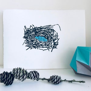 May include: A black and white linocut print of a bird's nest with blue eggs. The nest is made of twigs and branches and is surrounded by a white background.