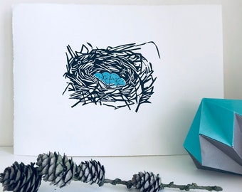 May include: A black and white linocut print of a bird's nest with blue eggs. The nest is made of twigs and branches and is surrounded by a white background.