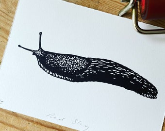 May include: Black and white linocut print of a slug on white paper. The print is titled "Red Slug" and signed by the artist.