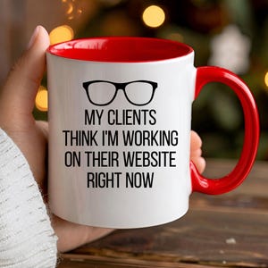 Könnte beinhalten: Ein weißer Keramikbecher mit leuchtend rotem Innenraum und Henkel. Auf der Vorderseite befindet sich eine schwarze Brillengrafik über dem Text: MY CLIENTS THINK I'M WORKING ON THEIR WEBSITE RIGHT NOW. Der Becher hat eine glatte Oberfläche und eine zylindrische Form.
