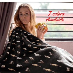 May include: A woman is sitting on a couch, wrapped in a black fleece blanket with a white ferret pattern. The blanket is soft and cozy. The text "7 colors available" is visible in the upper right corner of the image.