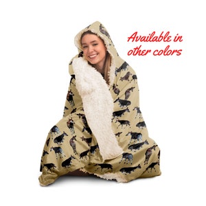 Op de afbeelding: Een hooded deken met een beige okapi-patroon en crème-kleurige voering. De tekst "Available in other colors" is rood zichtbaar. Ontworpen voor warmte en comfort.