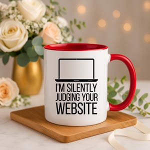 Könnte beinhalten: Weiße Keramiktasse mit rotem Rand und Henkel. Die Tasse zeigt eine schwarze Silhouette eines Laptops mit dem Text "I'm silently judging your website".