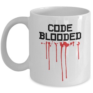 以下が含まれることがあります： 白いセラミック製のコーヒーカップ。黒い文字で「CODE BLOODED」と書いてあり、赤い血しぶきがカップから垂れている。