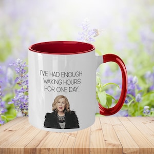 May include: White ceramic mug with a red interior and handle. The mug features the quote "I'VE HAD ENOUGH WAKING HOURS FOR ONE DAY." and an illustration of a woman. The mug is on a wooden surface with a blurred floral background.