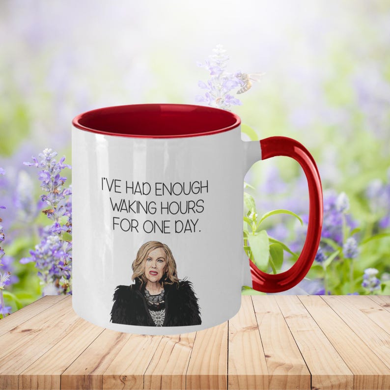 May include: White ceramic mug with a red interior and handle. The mug features the quote "I'VE HAD ENOUGH WAKING HOURS FOR ONE DAY." and an illustration of a woman. The mug is on a wooden surface with a blurred floral background.