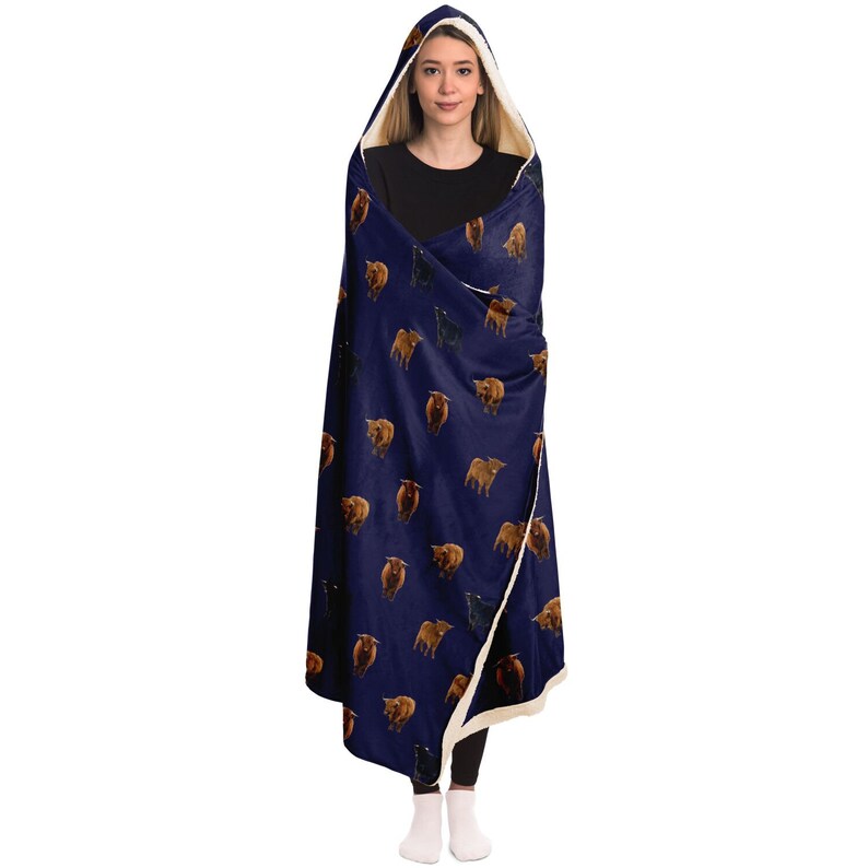 May include: A navy blue hooded blanket with a white sherpa lining. The blanket features a repeating pattern of brown and black Highland cattle.