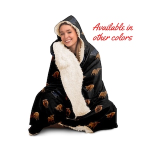 May include: A black hooded blanket with a white sherpa lining. The blanket features a repeating pattern of brown and black Highland cattle. The text "Available in other colors" is displayed in red.