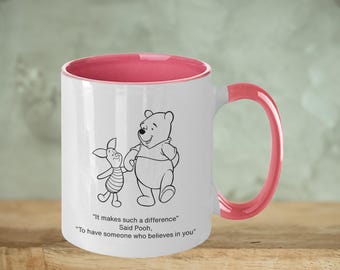 May include: White ceramic mug with a pink rim and handle. The mug features a black and white illustration of Winnie the Pooh and Piglet. The text on the mug reads: "It makes such a difference" Said Pooh, "To have someone who believes in you."