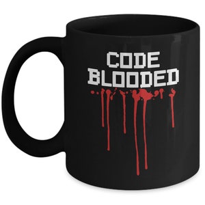 以下が含まれることがあります： 白いピクセル文字で「CODE BLOODED」と書かれた黒いセラミック製のコーヒーカップ。赤い血がカップから滴り落ちている。