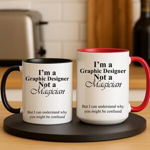 May include: Two white ceramic mugs with black and red accents. Each mug has the text "I'm a Graphic Designer Not a Magician" printed in black, along with the phrase "But I can understand why you might be confused."