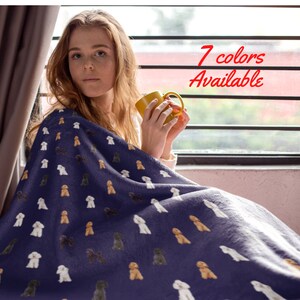 Pode incluir: Uma mulher está sentada perto de uma janela e segurando uma caneca amarela. Ela está enrolada em um cobertor de lã macio e roxo com um padrão repetido de poodles brancos, marrons e pretos. O texto "7 cores disponíveis" é exibido em letras vermelhas.