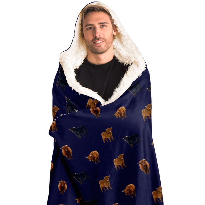 May include: A navy blue hooded blanket with a white sherpa lining. The blanket features a repeating pattern of brown and black Highland cattle.