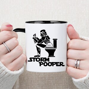 May include: White ceramic coffee mug with a black rim and handle. The mug features a black and white illustration of a Stormtrooper sitting on a toilet with the text "Storm Pooper".