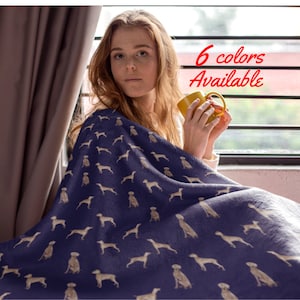 Puede incluir: Una mujer está sentada en un sofá con una taza de café en la mano. Está envuelta en una suave manta de franela azul con un patrón de perros beige. El texto "6 colores disponibles" está escrito en rojo sobre un fondo blanco.