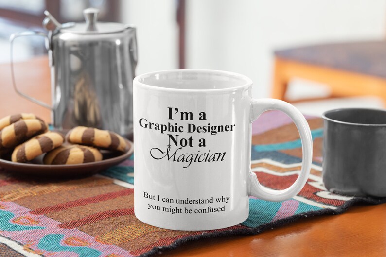 May include: White ceramic mug with the text "I'm a Graphic Designer Not a Magician" in black. The mug has a curved handle and is on a patterned surface. A teapot and cookies are in the background.