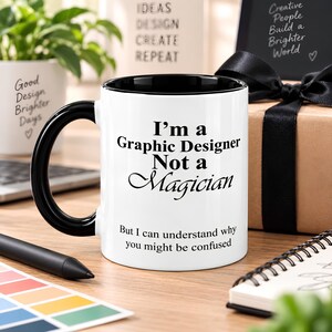Black accent graphic designer mug displayed beside a wrapped gift with subtle creative workspace details, styled as a gift for designers, freelancers, or creative coworkers.