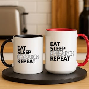 Può includere: Due tazze in ceramica bianca con dettagli neri e rossi. Ogni tazza presenta la scritta "EAT SLEEP RESEARCH REPEAT" in nero. Una tazza ha un manico e l'interno neri, l'altra ha un manico e l'interno rossi. Le tazze sono su un vassoio nero.