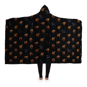 May include: A black hooded blanket with a repeating pattern of brown and black Highland cattle. The blanket is worn by a person standing on a white background.
