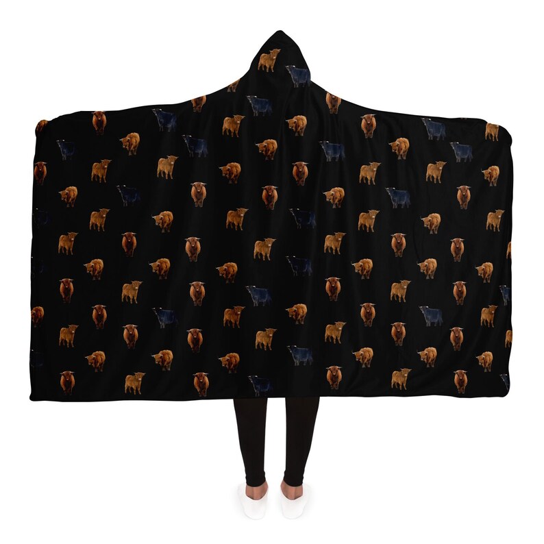 May include: A black hooded blanket with a repeating pattern of brown and black Highland cattle. The blanket is worn by a person standing on a white background.