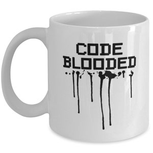 以下が含まれることがあります： 白いセラミック製のコーヒーカップで、黒い文字で「CODE BLOODED」と書いてあります。ピクセル化されたフォントで書かれています。黒い塗料が文字から垂れています。
