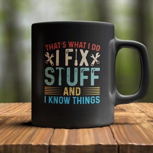 Op de afbeelding: Zwarte koffiemok met een vintage ontwerp. De tekst luidt "That's what I do I fix stuff and I know things".