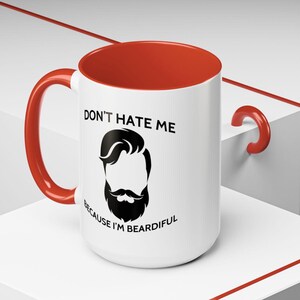 Puede incluir: Taza de cerámica blanca con borde y asa rojos. La taza tiene una ilustración en blanco y negro de un hombre con barba y el texto "No me odies porque tengo barba".