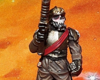 May include: A painted miniature figure of a soldier wearing a brown and black uniform with a red sash and a gas mask. The soldier is holding a rifle and standing on a snowy base.