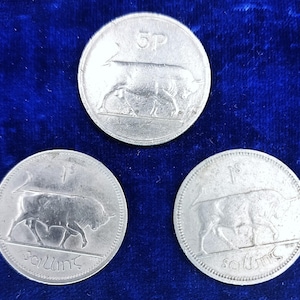 May include: Three silver coins with a bull design. One coin reads "5P" and the other two read "1S" and "Scilling". The coins are on a dark blue background.