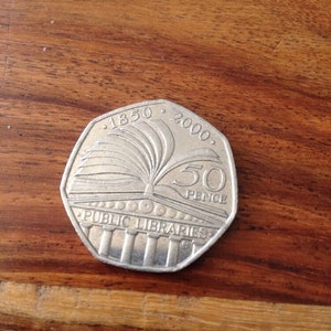 Puede incluir: Una moneda de 50 peniques de plata que conmemora el 2000 aniversario de las bibliotecas públicas en el Reino Unido. La moneda presenta un libro abierto con un árbol estilizado que crece de él, y el texto "1850 2000 PUBLIC LIBRARIES 50 PENCE".