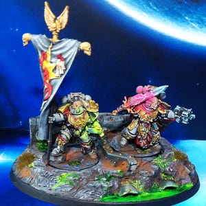 May include: Two painted miniature figures of space marines, one holding a sword and the other holding a gun, standing on a rocky base with a banner behind them.