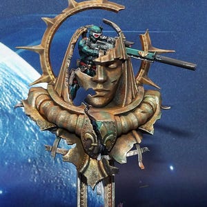 May include: A detailed miniature figure of a soldier aiming a rifle, set within a stylized, weathered bronze sculpture. The sculpture features a face, a halo-like structure, and ornate details. The color palette includes bronze, green, and touches of blue.