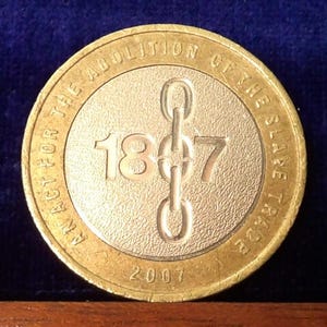 Puede incluir: Una moneda británica de dos libras de color dorado que conmemora la Ley de Abolición del Comercio de Esclavos de 1807. La moneda presenta un diseño de cadena estilizado y el año 2007.