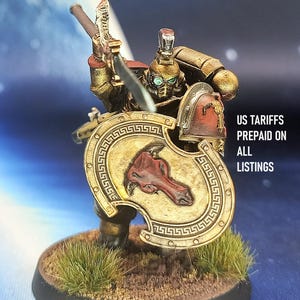Puede incluir: Figura en miniatura de un guerrero con armadura dorada y roja, sosteniendo una espada y un escudo. El escudo presenta un emblema rojo y un borde dorado. La base es redonda con hierba verde. El texto dice "US TARIFFS PREPAID ON ALL LISTINGS."