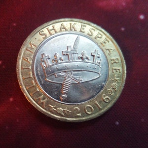 Pode incluir: Uma moeda dourada com um centro prateado que apresenta uma coroa com uma espada que a atravessa. A moeda diz "William Shakespeare" e "2016" ao redor da borda.