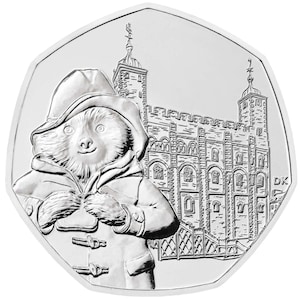 May include: A silver coin featuring Paddington Bear in a coat and hat, with the Tower of London in the background. The coin has a unique, irregular shape and is highly detailed, showcasing the bear's features and the architecture.
